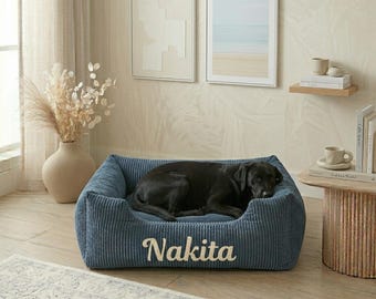 May include: A blue corduroy dog bed with the name "Nakita" embroidered on the front. A black dog is resting inside the bed. The bed is in a room with neutral-toned decor.