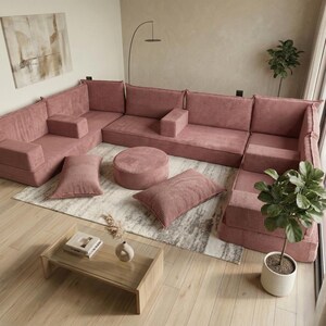 May include: A modular, U-shaped sectional sofa in a dusty rose corduroy fabric. The set includes multiple seat cushions, armrests, a round ottoman, and two rectangular throw pillows. The sofa sits on a patterned rug in a modern living room setting.
