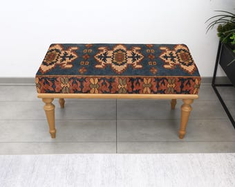Può includere: Un pouf rettangolare con un top a fantasia nei toni del blu, arancione e beige. Le gambe in legno sono marrone chiaro. Il pouf è un elemento d'arredo decorativo.