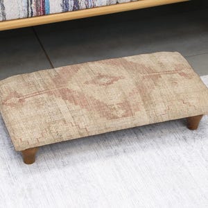 Op de afbeelding: Een rechthoekige voetenbank met een beige en rood patroon en vier korte, afgeronde houten poten. De voetenbank staat op een wit tapijt. De afmetingen zijn ongeveer 60 cm lang en 30 cm breed.