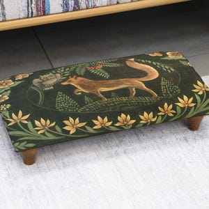 Puede incluir: Un reposapiés rectangular con una tela verde oscuro con una ilustración de un zorro. El zorro es marrón con una cola larga, rodeado de patrones florales y de hojas. El taburete tiene patas de madera y está colocado sobre una alfombra clara.