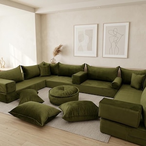 May include: A modular olive green velvet sectional sofa with multiple cushions and a round ottoman. The sofa is arranged in a U-shape, with several square and rectangular cushions scattered around. The room has light-colored walls and hardwood floors.