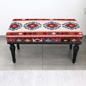May include: A rectangular ottoman with a patterned top and black legs. The top has a cream background with a repeating geometric design in red, blue, and brown. The legs are turned and painted black.