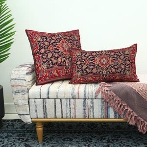 May include: Two decorative throw pillows with a floral pattern in red, navy, and cream colors. The pillows rest on a striped bench with a pink blanket and a potted plant in the background. The pillows have a traditional rug design.