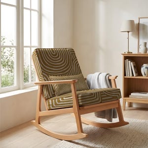 May include: A wooden rocking chair with a patterned seat and back. The fabric has an olive green and white circular design. A grey blanket is draped over the arm. The chair is in a bright room, next to a window and a bookshelf.