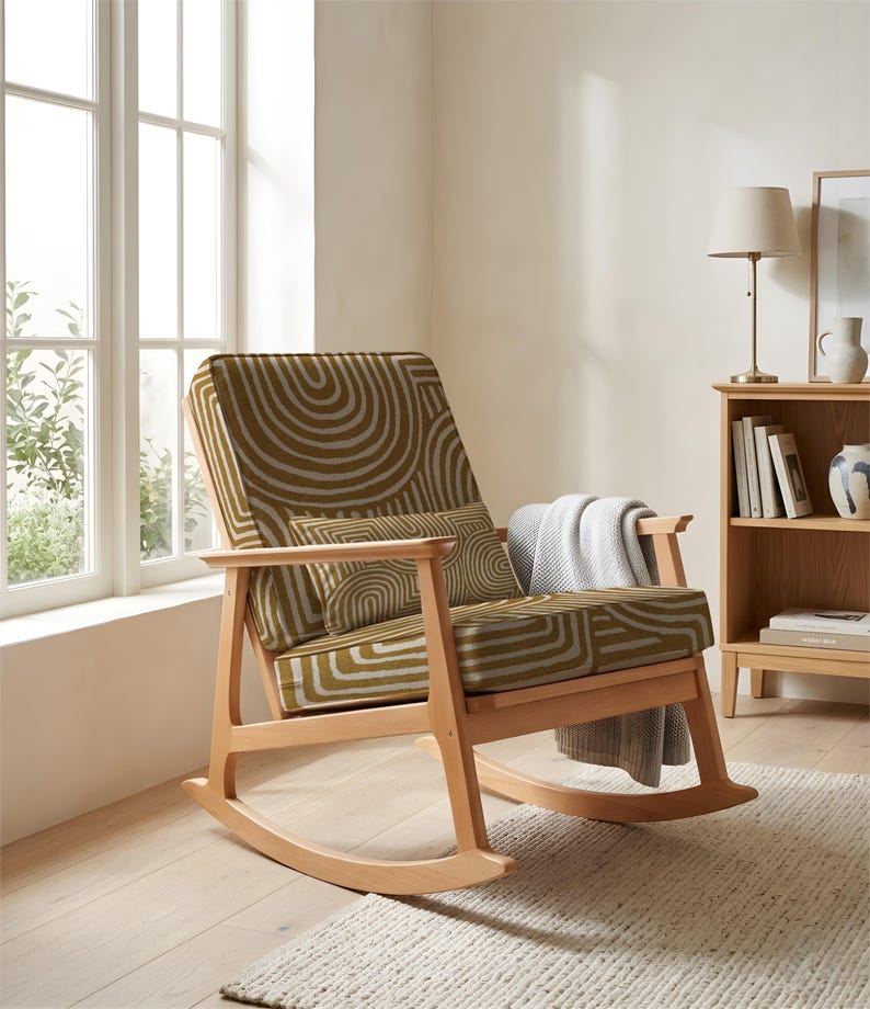 May include: A wooden rocking chair with a patterned seat and back. The fabric has an olive green and white circular design. A grey blanket is draped over the arm. The chair is in a bright room, next to a window and a bookshelf.