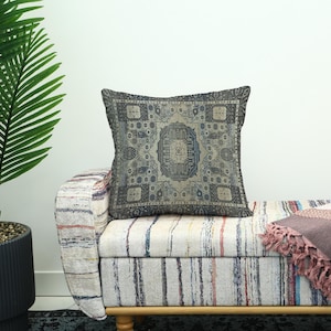 May include: A square cushion featuring a vintage rug pattern in blue, grey, and beige tones. The cushion rests on a striped bench with a wooden frame. A potted plant is visible on the left side of the image.