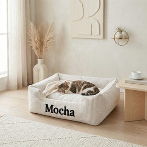 May include: A beige corduroy cat bed with the name "Mocha" embroidered in black. A brown and white tabby cat is curled up asleep on a cream-colored blanket inside the bed. The bed is rectangular and sits on a light wood floor.