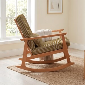 May include: A wooden rocking chair with a green and white geometric patterned cushion. The chair is in a bright room with a window, a small side table, and a plant. The chair has a classic design and is made of light-colored wood.