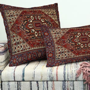 May include: Two decorative cushions with a traditional rug design in red, cream, and navy blue. The cushions are placed on a patterned sofa. The larger cushion is square, and the smaller one is rectangular.