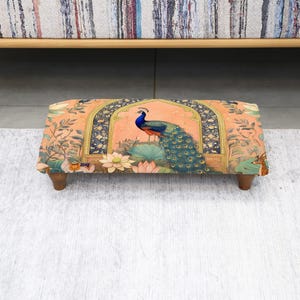 May include: A rectangular footstool with a vibrant peacock design. The fabric features a peach background with a peacock, lotus flowers, and ornate archways. The stool has four wooden legs and is placed on a light gray rug.