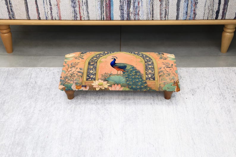 May include: A rectangular footstool with a vibrant peacock design. The fabric features a peach background with a peacock, lotus flowers, and ornate archways. The stool has four wooden legs and is placed on a light gray rug.