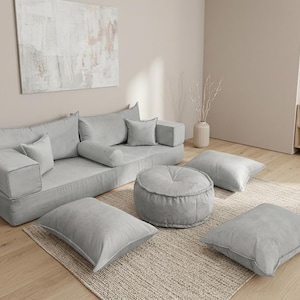 May include: A light grey modular sofa set with multiple cushions and a round ottoman. The set includes a large sofa, pillows, and floor cushions, all in a soft, neutral colour. The furniture is arranged on a neutral rug.