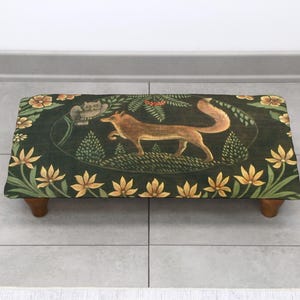 Op de afbeelding: Een rechthoekige voetenbank met een vossenmotief op een donkergroene achtergrond. De vos is bruin en beige, omringd door gele bloemen en groen gebladerte. De kruk heeft vier houten poten.