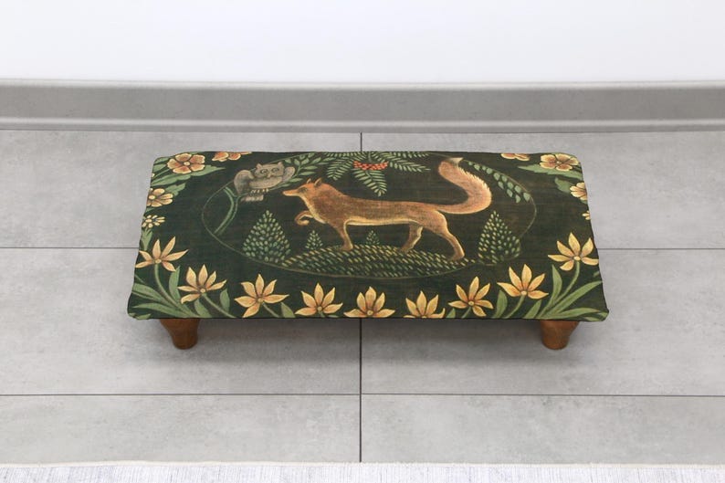 Op de afbeelding: Een rechthoekige voetenbank met een vossenmotief op een donkergroene achtergrond. De vos is bruin en beige, omringd door gele bloemen en groen gebladerte. De kruk heeft vier houten poten.