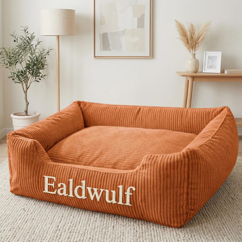 May include: A rectangular pet bed made of orange corduroy fabric with high, cushioned sides. The front panel features the name Ealdwulf embroidered in white, capitalized, serif lettering. The bed has a soft, plush interior cushion.