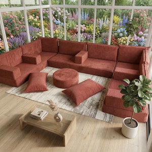 Puede incluir: Un sofá seccional modular de pana color óxido con otomana redonda y cojines a juego. Una mesa de centro con tapa de cristal y una planta en maceta están frente al sofá. La habitación tiene grandes ventanales con vistas a un jardín colorido.