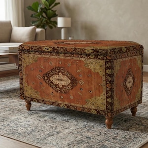 Può includere: Un pouf rettangolare con un tessuto fantasia marrone-rossastro. Il design presenta un medaglione centrale e accenti angolari in oro e marrone scuro. Il pouf poggia su quattro gambe corte in legno ed è posto su un tappeto fantasia.
