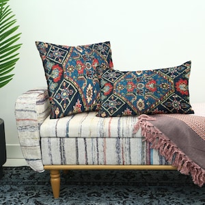 May include: Two decorative throw pillows with a dark blue, red, and gold geometric pattern rest on a striped bench. A pink blanket with fringe is draped over the bench. A potted palm tree is to the left.