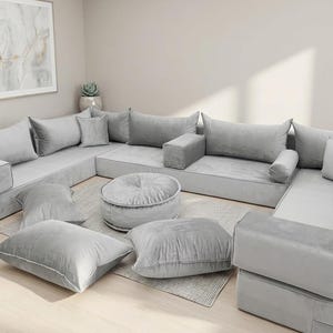 May include: A modular grey sectional sofa with multiple cushions and a round ottoman. The sofa is arranged in a U-shape, with several square and rectangular cushions. The ottoman and cushions are in a matching grey colour.