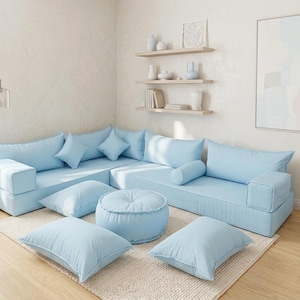 Puede incluir: Un sofá modular azul claro con cojines a juego y una otomana redonda. El sofá está dispuesto en forma de L sobre una alfombra beige. Estanterías decorativas y una obra de arte enmarcada son visibles.