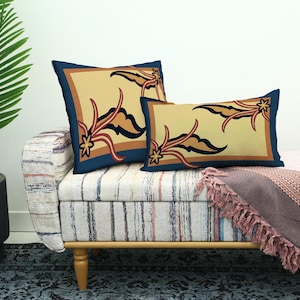 May include: Two decorative throw pillows with a beige background and a navy blue border. The pillows feature a stylized floral design in shades of brown, rust, and black. They rest on a striped bench with a pink throw blanket.