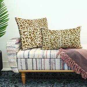 Könnte beinhalten: Zwei dekorative Kissen mit einem braun-weißen Fleckenmuster liegen auf einer gestreiften Bank. Eine rosa Decke mit Fransen ist über die Bank drapiert. Eine Topfpalme befindet sich im Hintergrund.