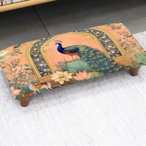 May include: Rectangular footstool with a vibrant peacock design. The fabric features a peach background with a peacock, lotus flowers, and ornate archways. The stool has dark wooden legs and is placed on a light-colored rug.