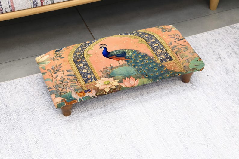 May include: Rectangular footstool with a vibrant peacock design. The fabric features a peach background with a peacock, lotus flowers, and ornate archways. The stool has dark wooden legs and is placed on a light-colored rug.