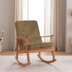 May include: A wooden rocking chair with a light brown frame and a seat and backrest upholstered in a green and white geometric pattern. The chair is placed on a white rug in front of a window.