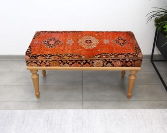 May include: A rectangular ottoman with a vibrant, patterned rug top in shades of red, orange, and brown. The ottoman sits on four light-colored wooden legs. The overall design is a blend of traditional rug artistry and modern furniture.