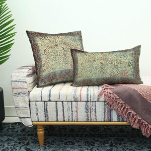 May include: Two decorative throw pillows with a floral pattern in shades of green, brown, and red rest on a striped bench. A matching patterned blanket with fringe is draped over the bench. A potted plant is visible on the left.