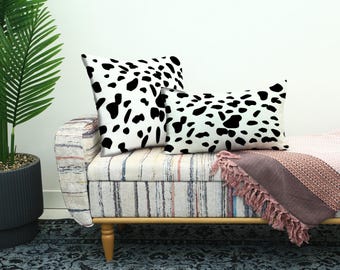 May include: Two decorative throw pillows with a white background and black dalmatian-like spots. The pillows are on a striped bench with a pink blanket. A potted palm tree is on the left.