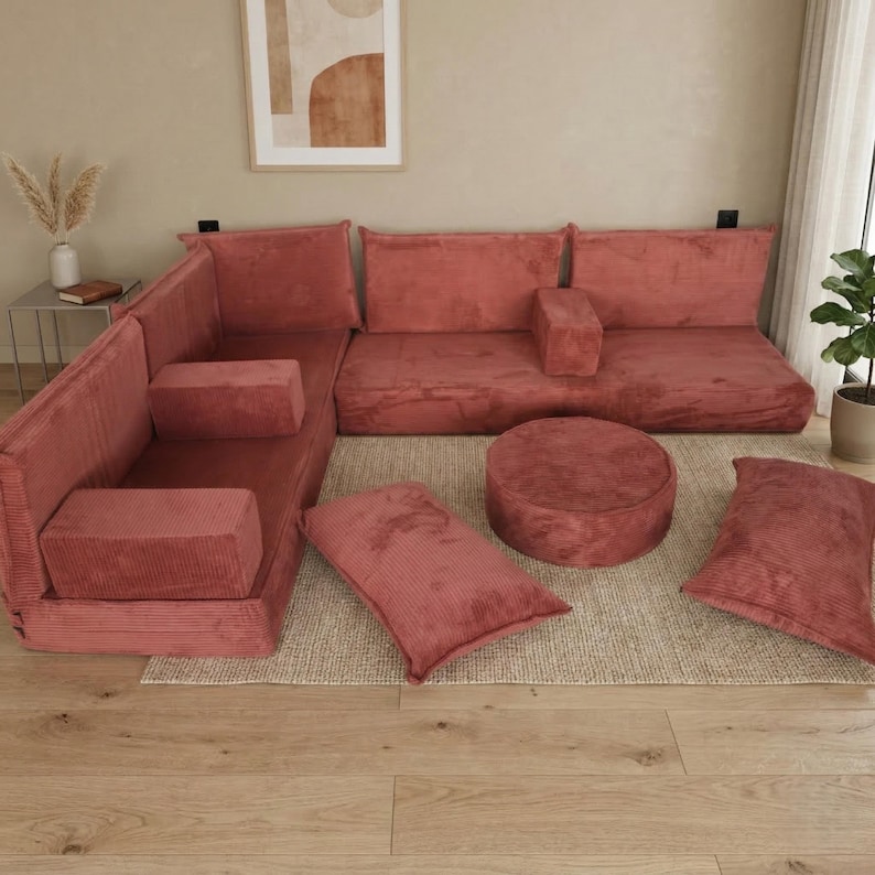 Op de afbeelding: Een modulaire, koraalkleurige corduroy hoekbank met meerdere kussens en een ronde poef. De bank is geplaatst op een neutraal getint tapijt, waardoor een gezellige en uitnodigende woonruimte ontstaat. Het ontwerp is modern en veelzijdig.