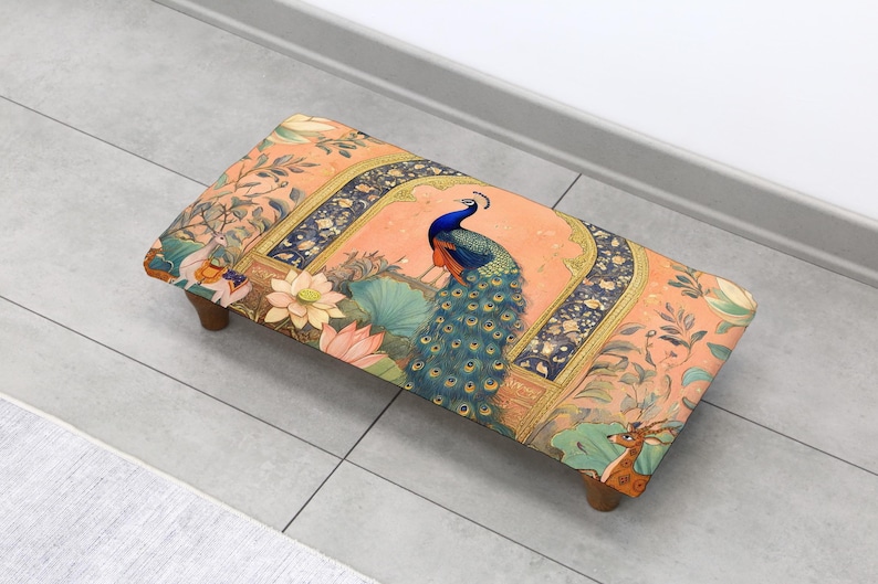May include: A rectangular footstool with a decorative peacock design. The stool features a peach-colored background with a peacock, lotus flowers, and a deer. The legs are made of wood.
