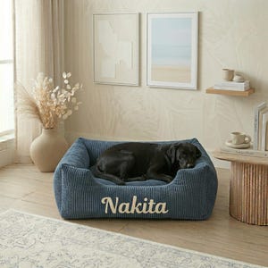 May include: A rectangular, navy blue corduroy pet bed with raised sides and a soft interior. The front of the bed features the name Nakita embroidered in a cream-colored, cursive script font. A black dog is lying inside the pet bed.