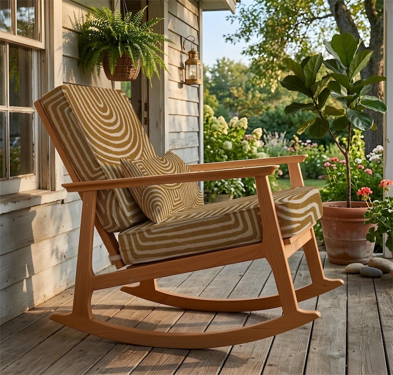 May include: A wooden rocking chair with a light brown finish. The seat and back cushions have a repeating pattern of concentric, curved lines in tan and cream. The chair is on a wooden porch, with plants and a hanging lantern in the background.
