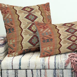 May include: Two decorative cushions with a geometric pattern in beige, red, and teal. The cushions are placed on a patterned sofa. The design features diamond and zigzag motifs, adding a touch of Southwestern style.