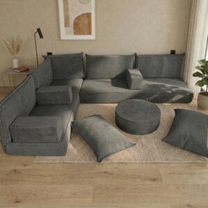 May include: A modular gray corduroy sofa arrangement with multiple sections, including corner pieces, a central seat, and an ottoman. Several matching rectangular and round cushions are arranged on a neutral rug, creating a comfortable seating area.