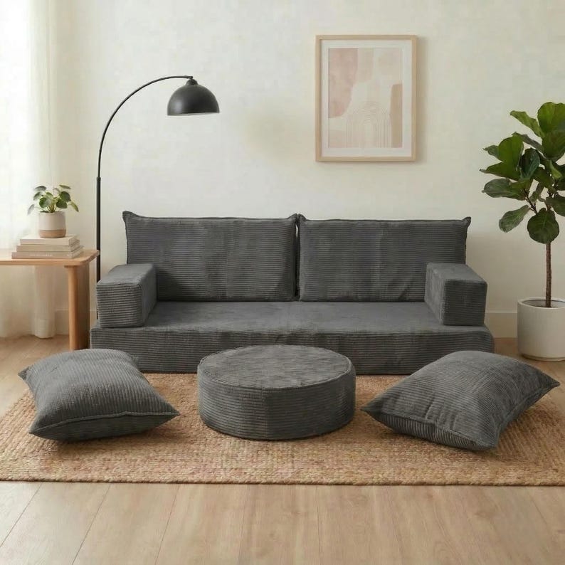 Op de afbeelding: Een grijze modulaire bankset van corduroy met twee rugkussens, twee armleuningen, een ronde poef en twee vierkante kussens. De meubels staan op een geweven vloerkleed in een lichte kamer. Een vloerlamp en een potplant zijn ook zichtbaar.