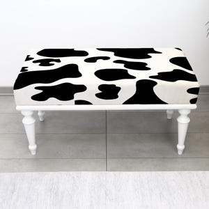 May include: A rectangular bench with a white frame and legs. The cushioned top features a black and white cow print pattern. The bench is placed on a gray tiled floor, with a white wall in the background. A black planter with greenery is visible.