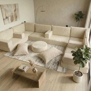 May include: A modular beige corduroy sectional sofa with matching pillows and a round ottoman. The sofa is arranged in an L-shape on a patterned rug, with a wooden coffee table and potted plants nearby. The room has a neutral color palette.