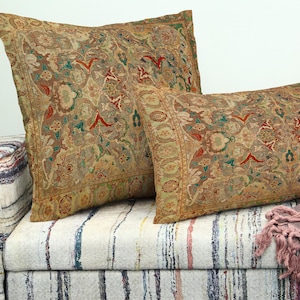 May include: Two decorative cushions with an ornate, vintage-style pattern in shades of beige, green, and red. The cushions rest on a striped upholstered sofa, creating a cosy and inviting aesthetic. The cushions have a rectangular and square shape.