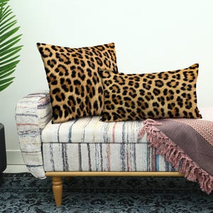 Puede incluir: Dos cojines con estampado de leopardo descansan sobre un banco a rayas con una manta rosa. Los cojines tienen un fondo beige con manchas marrones oscuras. El banco tiene patas de madera y una tela estampada.