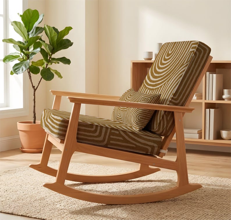 May include: A wooden rocking chair with a light brown frame and olive green and white patterned upholstery. The chair is set on a beige rug. A potted plant and a wooden shelf are visible in the background. The chair's design is modern.