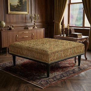Pode incluir: Um otomano quadrado com tecido estampado multicolorido, apresentando motivos florais e geométricos complexos em tons de dourado, ferrugem e azul-petróleo. A peça é sustentada por quatro pernas cónicas de madeira castanho-escura.