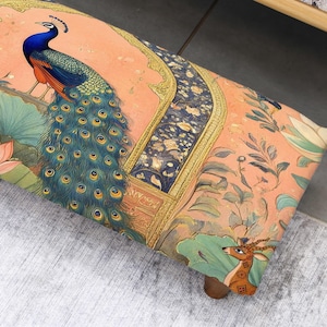 May include: A decorative ottoman with a vibrant peacock design. The ottoman features a peach background with a detailed illustration of a peacock, lotus flowers, and a deer. The edges are adorned with gold trim.