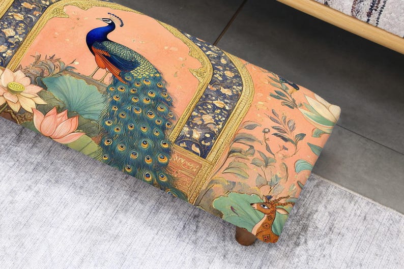 May include: A decorative ottoman with a vibrant peacock design. The ottoman features a peach background with a detailed illustration of a peacock, lotus flowers, and a deer. The edges are adorned with gold trim.