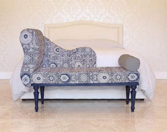 May include: A chaise lounge with a blue and gray patterned fabric, set against a white bed and a patterned wall. The chaise has a curved back and a rectangular seat, supported by dark blue legs. A gray bolster pillow rests on the seat.