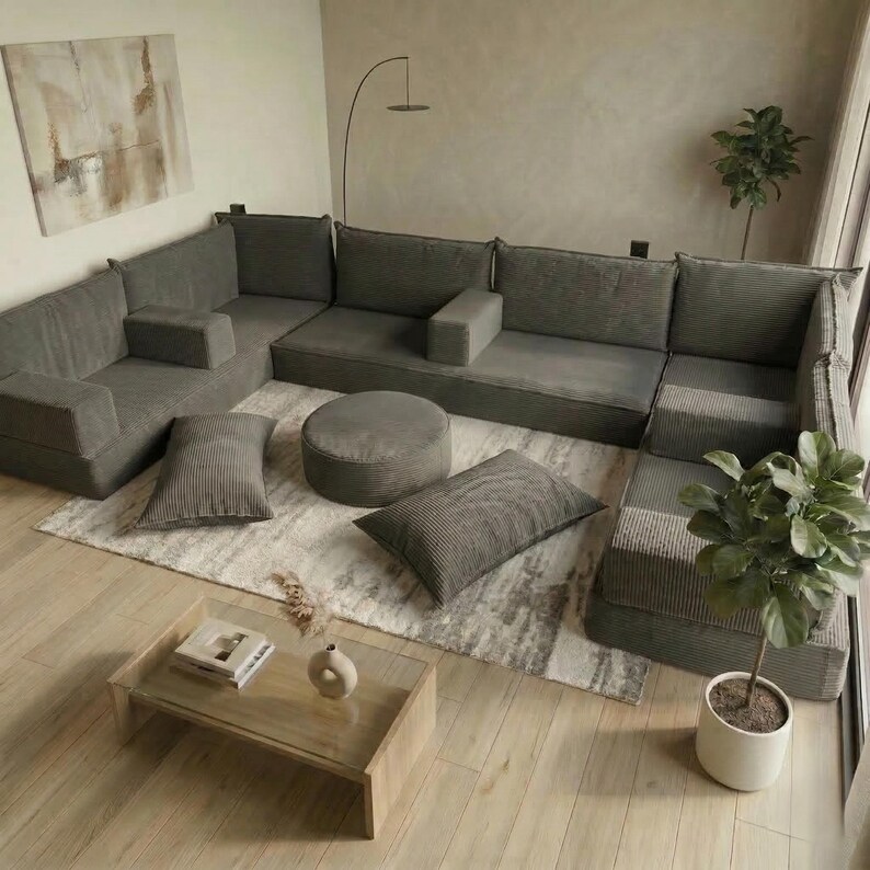 Op de afbeelding: Een modulaire grijze hoekbank met bijpassende kussens en een ronde poef. De bank is in een U-vorm op een lichtgekleurd tapijt geplaatst, met een houten salontafel en een plant in pot in de buurt. De kamer heeft een neutraal kleurenpalet.